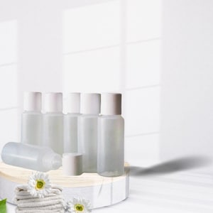 May include: Six clear plastic bottles with white caps on a wooden platform. The bottles are arranged in a row, and there is a white towel and two white flowers in front of them.