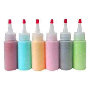 Può includere: Sei piccole bottiglie di sabbia colorata, ciascuna con un tappo rosso. Le bottiglie sono disposte in fila da sinistra a destra: rosa, blu, arancione, rosa chiaro, grigio e verde.