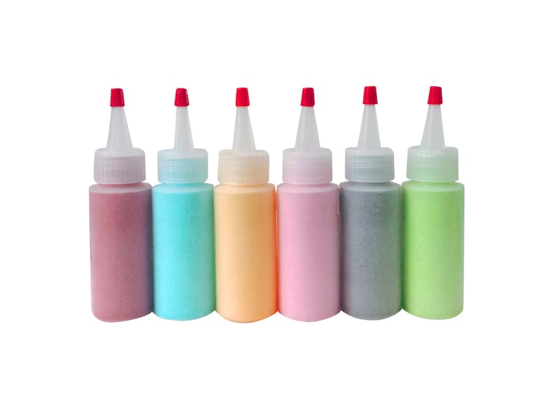 May include: Six small bottles of coloured sand, each with a red cap. The bottles are arranged in a row from left to right: pink, blue, orange, light pink, grey, and green.