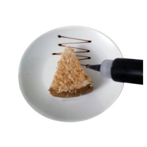 May include: A white plate with a slice of coconut cake and a black squeeze bottle with chocolate icing. The icing is being squeezed onto the plate to create a design.