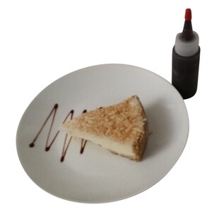 May include: A slice of coconut cake on a white plate with a chocolate drizzle. A small bottle of chocolate sauce is in the background.