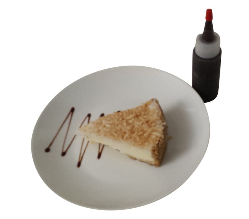May include: A slice of coconut cake on a white plate with a chocolate drizzle. A small bottle of chocolate sauce is in the background.