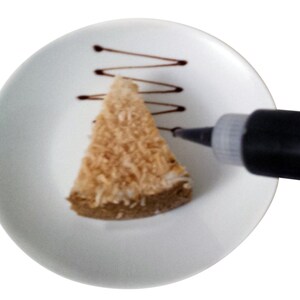 May include: A white plate with a slice of coconut cake on it. A black squeeze bottle is being used to drizzle chocolate over the cake.