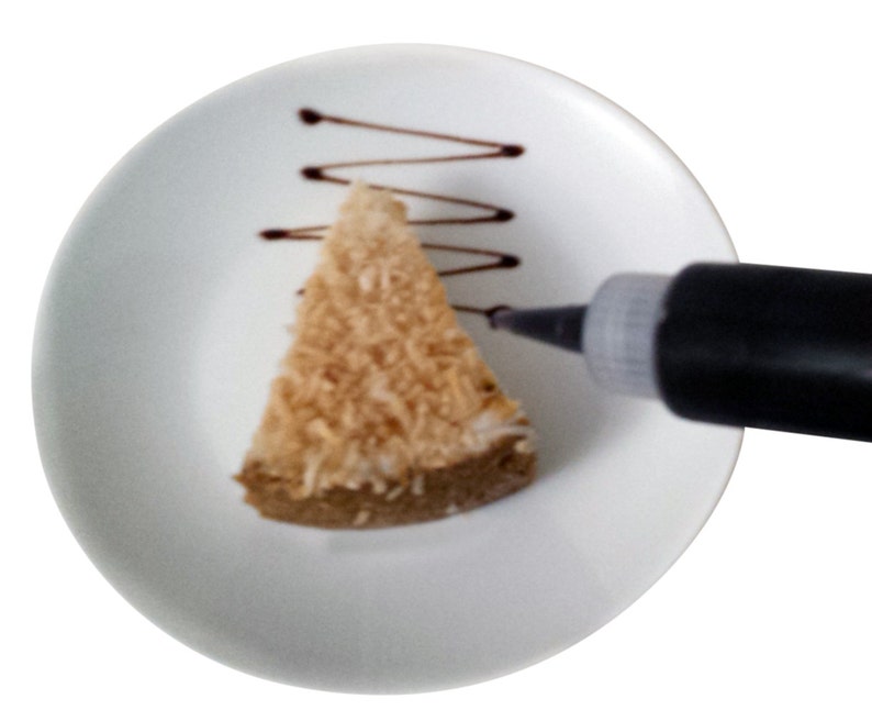May include: A white plate with a slice of coconut cake on it. A black squeeze bottle is being used to drizzle chocolate over the cake.