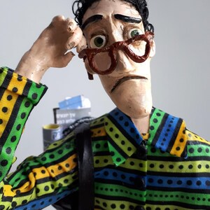 Peut inclure: Une figurine artisanale représentant une personne aux cheveux noirs bouclés, portant des lunettes à monture brune et une chemise à col à rayures horizontales jaunes, vertes et bleues avec des pois noirs. La figurine a une expression soucieuse.