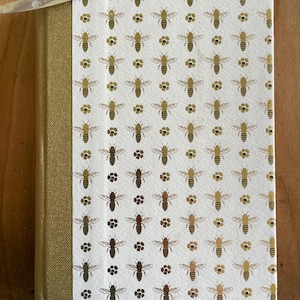 May include: A hardcover book with a gold fabric spine and a white cover featuring a repeating pattern of gold foil bees and small gold flower icons. A thin metal tool is visible at the top left corner of the book cover, resting on the edge.