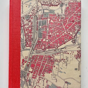 May include: A rectangular hardcover book featuring a bright red fabric spine and a front cover printed with a detailed vintage-style map. The map displays urban street layouts in shades of cream, grey, and pink, with various landmarks labeled.