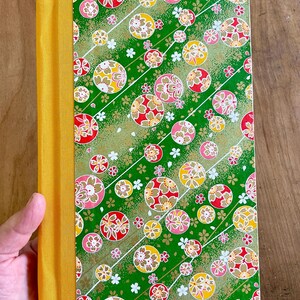 May include: A green journal with a yellow spine and a colorful Japanese-inspired pattern of circles and flowers. The cover is textured and has a subtle shimmer.