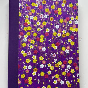 May include: A rectangular notebook featuring a solid purple fabric spine and a front cover decorated with a vibrant purple background, gold swirling line patterns, and a dense print of small yellow, white, and pink five-petaled flowers.