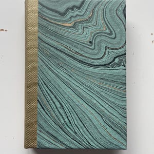 May include: A hardcover book featuring a textured gold fabric spine and a cover with a marbled pattern in shades of sage green, black, and metallic copper. The swirling, fluid lines create a topographical effect across the rectangular surface.