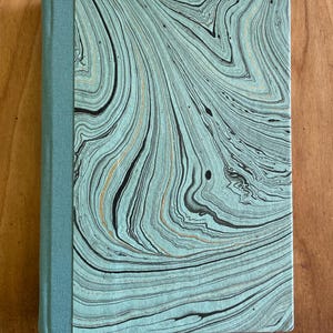 May include: A hardbound journal with a teal fabric spine and a cover featuring a swirling pattern in shades of teal, black, and gold. The notebook is rectangular and appears to be closed, resting on a wooden surface.