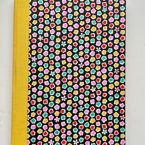 May include: A hardcover book featuring a solid yellow fabric spine and a black cover decorated with a repeating pattern of small, multi-colored flower shapes in shades of red, pink, yellow, and teal, arranged in a dense, uniform grid layout.