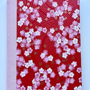 May include: A hardcover book featuring a vibrant red cover with a repeating pattern of pink and white cherry blossoms and subtle gold accents. The spine is bound in a solid light pink fabric, providing a contrast to the floral design.