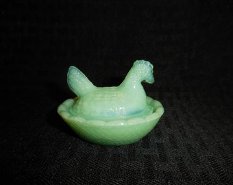 May include: A light green glass hen-on-nest figurine. The hen is perched atop a matching nest, with a textured surface and a slightly iridescent finish. The figurine is a decorative item.