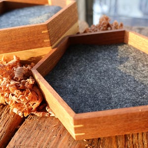 Puede incluir: Dos bandejas hexagonales de madera con forro de fieltro gris. Las bandejas están apiladas una encima de la otra y están rodeadas de virutas de madera.