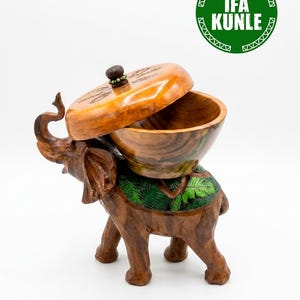 Pode incluir: Uma figura de elefante em madeira esculpida à mão com uma taça nas costas e uma tampa removível. O elefante tem veios de madeira castanha, detalhes de folhas pintadas a verde nas costas e um pequeno botão escuro com contas. Texto IFA KUNLE.