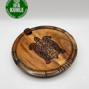 Puede incluir: Una bandeja redonda de madera con un diseño de tortuga quemado en color marrón oscuro en la superficie. El borde de la bandeja tiene detalles de líneas talladas y una pequeña cara esculpida en el borde. El logo verde IFA KUNLE es visible.