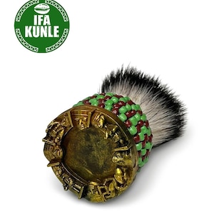 Puede incluir: Una brocha de afeitar con base dorada y relieves de figuras humanas. El mango está envuelto en un patrón de cuentas verdes y granates. El cabezal tiene cerdas densas en blanco y negro. Un logotipo verde muestra el texto IFA KUNLE.