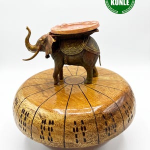 Pode incluir: Uma tigela de madeira polida com formato arredondado e marcas escuras gravadas nas laterais. Uma estatueta de elefante de metal com uma sela ornamentada e uma peça superior plana e circular repousa sobre a tampa. Um logótipo verde diz IFA KUNLE.