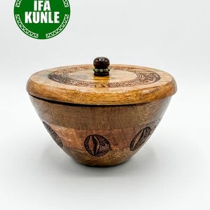 Puede incluir: Un cuenco redondo de madera con tapa a juego, que presenta un pomo de color marrón oscuro con pequeñas cuentas verdes. El exterior del cuenco está decorado con patrones grabados de conchas de cauri. Se ve un logotipo verde con IFA KUNLE.