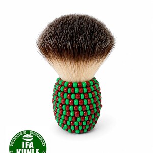 Puede incluir: Una brocha de afeitar con un cabezal de cerdas marrón oscuro y marrón claro. El mango está decorado con cuentas rojas y verdes. El cepillo tiene un logotipo verde redondo con el texto "IFA KUNLE".