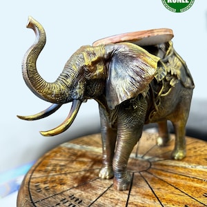 May include: A bronze-toned elephant figurine with a raised trunk and tusks, featuring intricate skin texture and leaf-like details on its back. It stands on a round wooden base with engraved radial lines and markings. A green circular logo reads IFA KÜNLE.