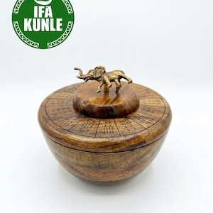Puede incluir: Un cuenco redondo de madera con un acabado marrón pulido y una tapa a juego. La tapa presenta una figura de elefante color latón como asa y está decorada con líneas radiales grabadas. Se ve un logotipo verde con el texto IFA KUNLE.