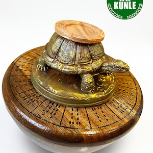Puede incluir: Una figura de tortuga de metal en tono dorado con un caparazón texturizado se asienta sobre una base circular de madera con líneas radiales y marcas grabadas. Un pequeño disco circular de color marrón claro descansa sobre el lomo. Un logo verde dice IFA KUNLE.