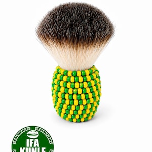 Puede incluir: Una brocha de afeitar con un mango cubierto por un patrón de pequeñas cuentas amarillas y verdes. El cabezal de la brocha presenta cerdas densas de color marrón oscuro que cambian a un tono beige claro en la base. Incluye un logotipo circular verde con el texto IFA KUNLE.
