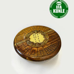 Puede incluir: Un cuenco redondo de madera con un acabado pulido en color marrón oscuro. La tapa presenta una incrustación circular central de latón con arena dorada texturizada y símbolos grabados, rodeada por un patrón radial de líneas negras.