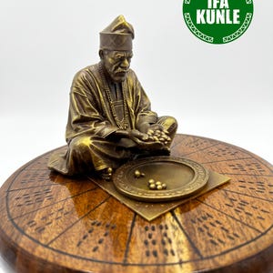 Pode incluir: Uma estatueta cor de bronze de uma pessoa sentada com um gorro e túnica tradicionais, segurando pequenas esferas sobre uma bandeja circular. A figura repousa sobre uma base de madeira polida com marcas gravadas e o texto IFA KUNLE.