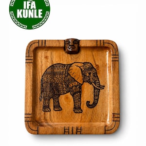 Puede incluir: Una bandeja cuadrada de madera con una ilustración de elefante pirograbada con intrincados patrones geométricos en el cuerpo. La bandeja tiene un detalle de cara tallada en el borde superior y marcas lineales a lo largo del borde. Un logotipo circular verde con el texto IFA KUNLE es visible en la esquina superior izquierda.