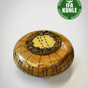 Puede incluir: Un cuenco redondo de madera con acabado pulido, que presenta marcas verticales negras grabadas en el exterior. La tapa central es una placa metálica circular de tono bronce con figuras en relieve y un centro texturizado. Un logotipo verde dice IFA KUNLE.