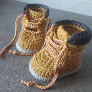 May include: Mustard yellow crochet baby boots, styled like work boots. They feature grey cuffs, white soles, and brown and pink laces. The handmade boots are crafted with a textured crochet stitch, offering a unique look.