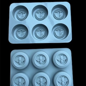 May include: Two pale blue silicone moulds, each with six circular cavities. Each cavity features a raised bee design. The moulds are designed for crafting soaps, candles, or other small items. The background is black.