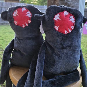 Może przedstawiać: Dwie pluszowe zabawki w kształcie stworzenia z ciemnoszarymi ciałami, długimi kończynami i dużymi czerwono-białymi plamami przypominającymi oczy. Siedzą na drewnianym stołku. Zabawki mają miękką, puszystą fakturę.