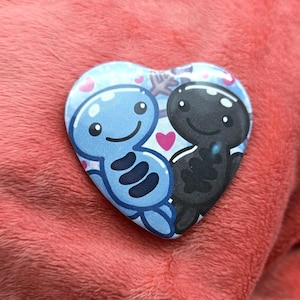 Puede incluir: Botón en forma de corazón con dos personajes de dibujos animados, uno azul y otro negro, abrazándose. La figura azul tiene tres rayas azul oscuro y la figura negra tiene un diseño texturizado. Corazones rosas y copos de nieve decoran el fondo.