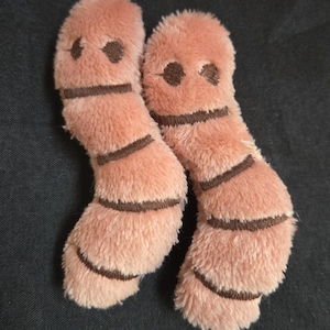 May include: Two plush, worm-shaped toys in a soft peach color. Each worm has dark brown stripes and two dark brown dots for eyes. The toys are made of a fuzzy material.