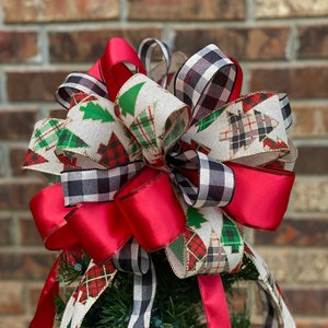May include: A festive Christmas tree bow featuring a mix of red, black and white plaid, and patterned ribbons with green Christmas trees. The bow is atop a small artificial tree, perfect for holiday decor.