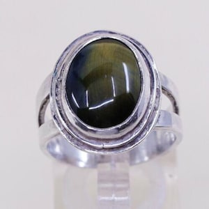 May include: Oval-shaped ring with a large, polished, olive-green gemstone set in a silver-toned band. The ring has a split shank design and a textured bezel around the stone. The gemstone has a chatoyant effect.