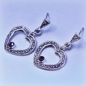 May include: A pair of silver-toned heart-shaped earrings. Each earring features a heart outline studded with small, dark stones and a single, larger red stone at the heart's center. The earrings have a decorative top piece and a hook closure.