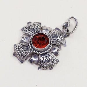 May include: A silver-toned pendant with a central, faceted, round, red gemstone. The pendant is shaped like a flower with detailed, textured petals. The bail is a simple loop for a chain. The pendant is likely vintage or antique.