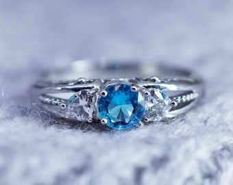 May include: A silver ring featuring a central, round, vibrant blue gemstone, with two smaller, clear, triangular stones either side. The band has a detailed, textured pattern.