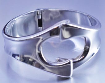Puede incluir: Una pulsera plateada pulida con un diseño abierto único. La pulsera presenta una forma curva con un recorte central y un cierre de bisagra. El metal refleja la luz, creando una apariencia brillante. Una pieza de joyería moderna.