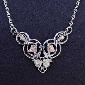 May include: Silver-tone necklace with a leaf design. The necklace features a decorative pendant with silver and gold-tone leaves. The chain is a delicate silver color, and the pendant has an intricate, openwork design.