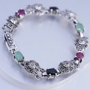 May include: Silver bracelet featuring alternating floral designs and colorful gemstones. The bracelet includes oval-shaped green, red, and black stones. The clasp is rectangular with a marcasite design.