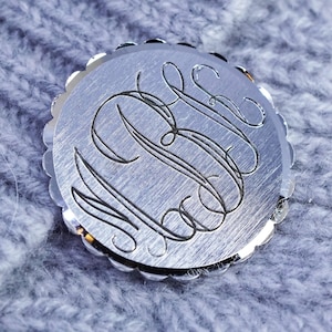 Puede incluir: Un colgante redondo plateado con monograma y borde festoneado. El colgante presenta un monograma grabado en escritura elegante. El fondo es una tela texturizada azul claro.