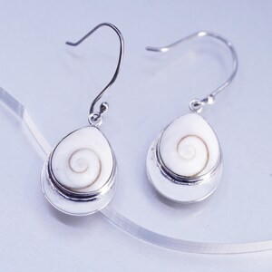 Könnte beinhalten: Ein Paar Ohrhänger mit weißen, tropfenförmigen Shiva-Auge-Muscheln, die ein natürliches braunes Spiralmuster aufweisen. Jede Muschel ist in eine silberfarbene Metallfassung eingefasst und mit einem passenden silberfarbenen Haken versehen.