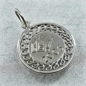 May include: A silver-colored pendant with a circular design. The pendant features an outer filigree border and a central disc with embossed text and symbols. The year "2006" is visible on the disc. The pendant has a small loop for a chain.