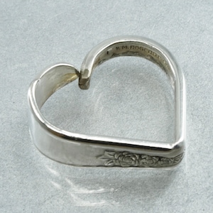 May include: A silver heart-shaped ring made from a spoon. The ring has a floral design on the side and the text "W.M. ROGERS MFG. CO." is visible. The ring is a unique piece of jewelry.